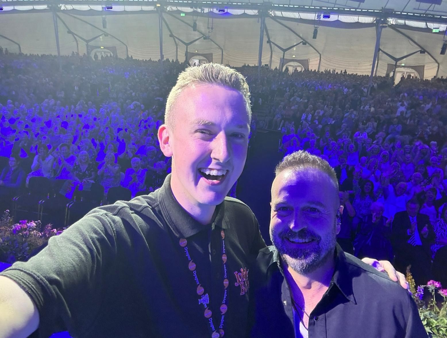 New trustee Shea Ferron on stage at Llangollen2023 with Alfie Boe, taking a selfie showing the audience in the background.